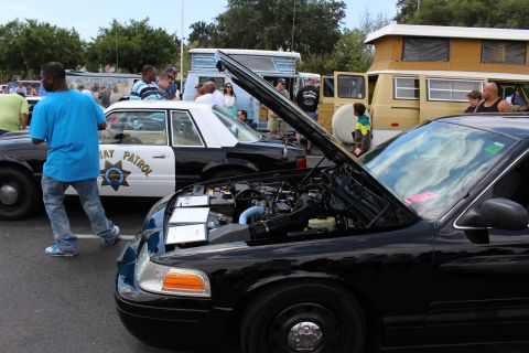 2003 Ford Crown Victoria - black sedan with its hood raised sits beside a Highway Patrol-marked car at a crowded outdoor show.