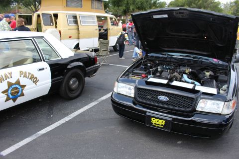 2003 Ford Crown Victoria - black Ford sedan displays its open hood and engine bay beside a black-and-white Highway Patrol car at a show.