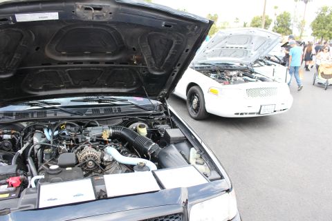 2003 Ford Crown Victoria - open engine bay of a dark sedan in the foreground, with a white sedan behind it, both hoods raised at a car show.