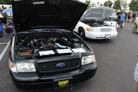 2003 Ford Crown Victoria - black Ford sedan with hood up reveals its engine, with a white sedan also hood-up behind it at a car show.