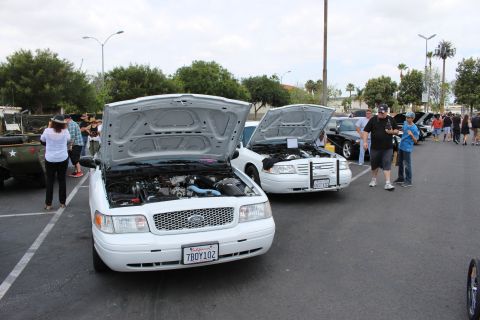 2003 Ford Crown Victoria - Two white sedans sit with hoods raised at a car show, the nearer one showing a California plate reading 7BOY102.
