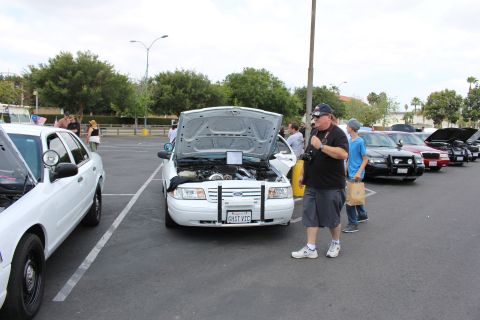 2003 Ford Crown Victoria - man in a cap stands beside a white Ford sedan with its hood raised among a row of cars at a parking-lot show.