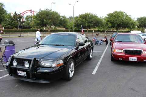 2003 Ford Crown Victoria - black sedan with a brush guard is parked next to a red Mercury sedan at an outdoor car meet with a roller coaster behind.