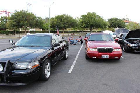 2003 Ford Crown Victoria - black sedan and a red Mercury sedan sit side by side in a parking lot with trees and a coaster in the background.
