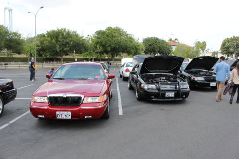 2003 Ford Crown Victoria - red Mercury Grand Marquis is parked next to black sedans with raised hoods at a parking-lot car show.