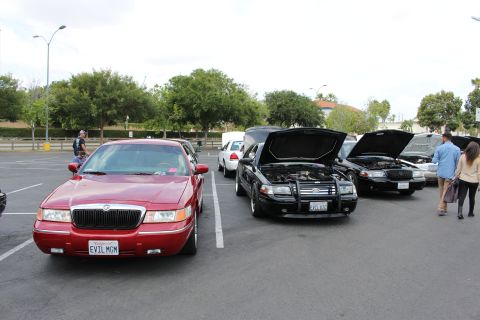 2003 Ford Crown Victoria - red Mercury sedan sits beside black sedans displaying open hoods at an outdoor parking-lot meet.