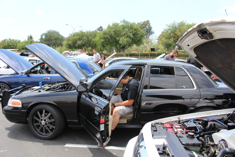 Black Ford Crown Victoria with hood up and a person seated in the open driver doorway at a car show