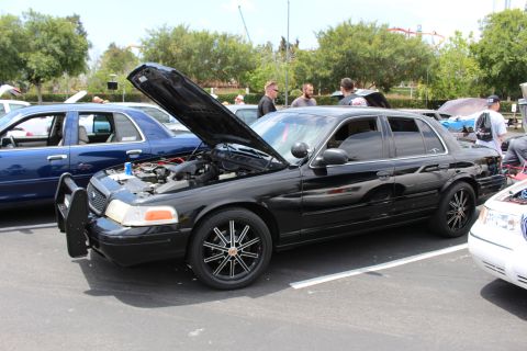 Black Ford Crown Victoria with raised hood, push bar and black multi-spoke wheels at a car show