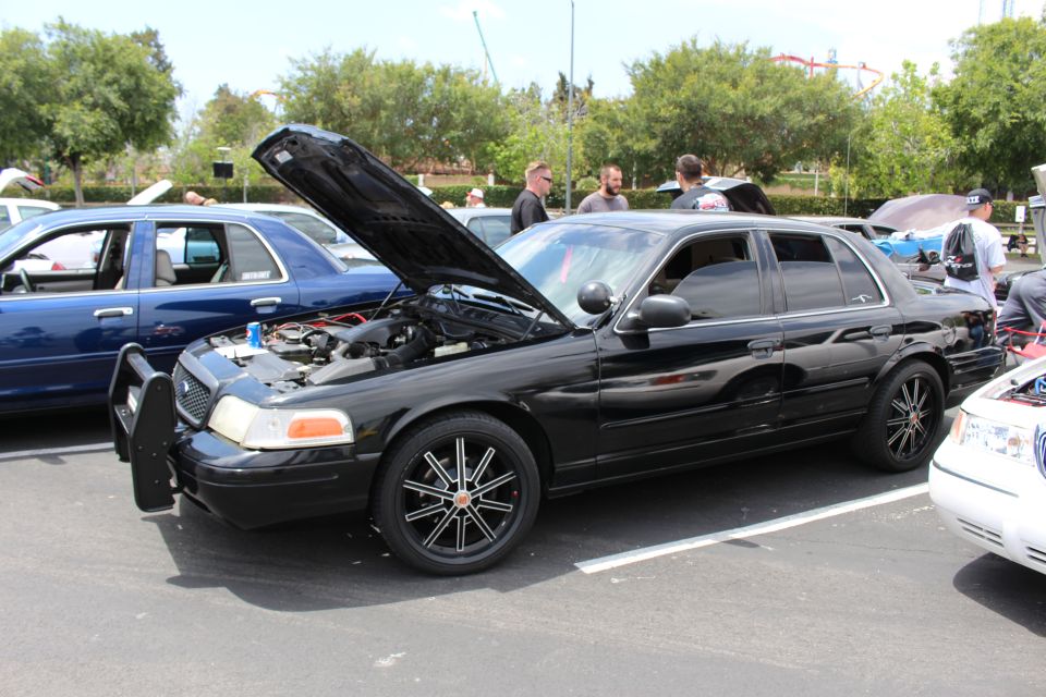 Black Ford Crown Victoria with raised hood, push bar and black multi-spoke wheels at a car show