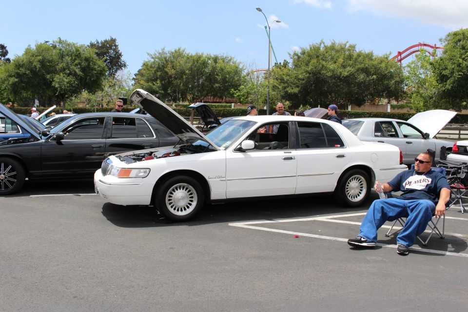 White Mercury Grand Marquis with hood up and a man seated in a chair beside it at a car show