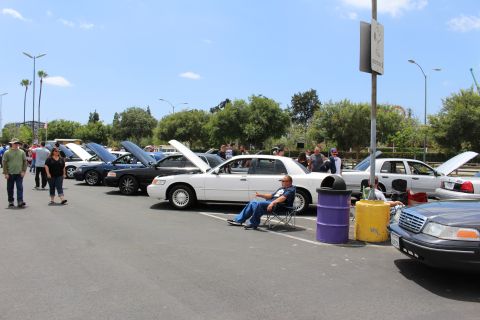 Row of full-size sedans with open hoods at a car show, a man seated in a chair and a purple barrel