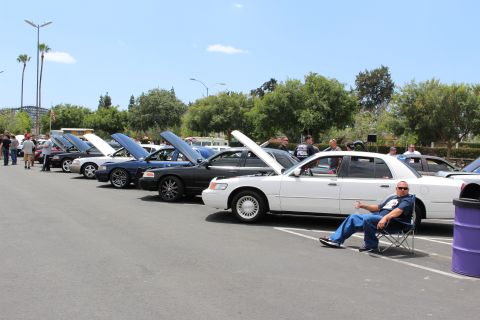 White Mercury Grand Marquis at the end of a row of open-hood sedans, man seated in a chair