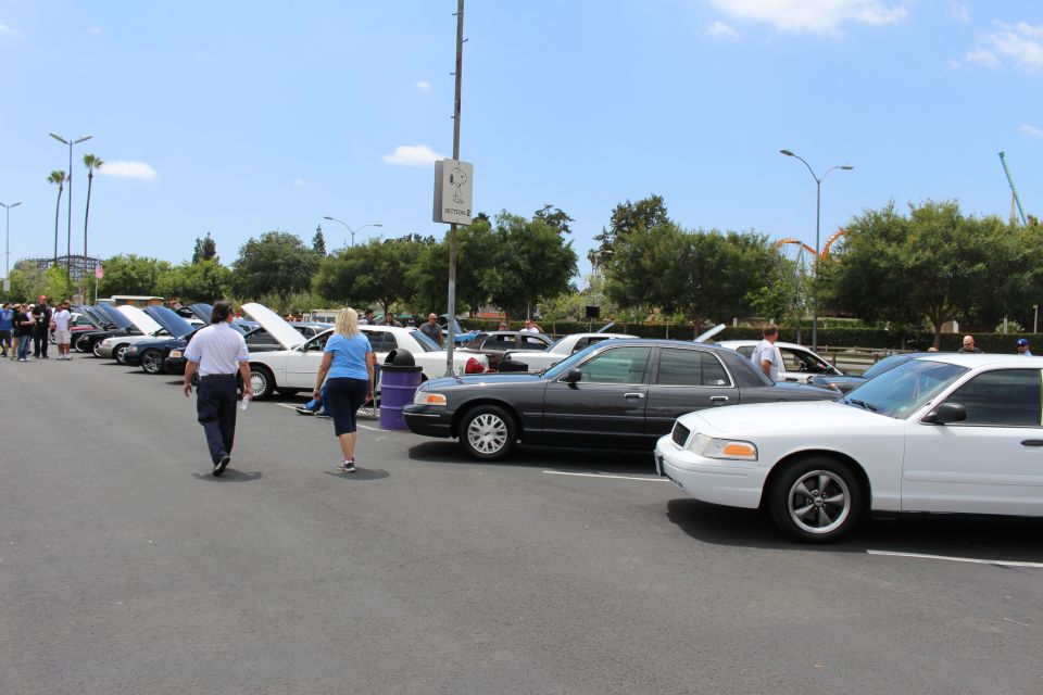 People walking through a parking lot of full-size sedans with open hoods at a car show