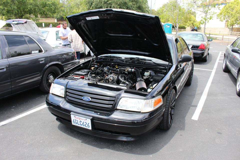 Black Ford sedan with hood open showing engine bay at a parking-lot car meet