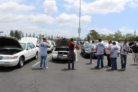 Crowd of people in matching gray T-shirts standing among parked sedans at a car meet