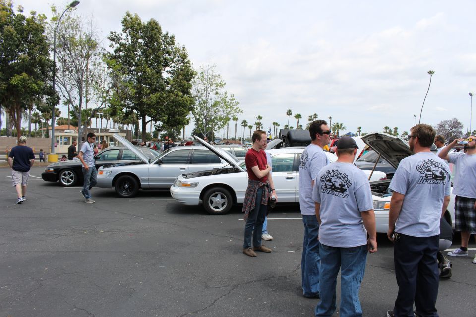 People in gray T-shirts standing beside silver and white sedans with open hoods at a car meet