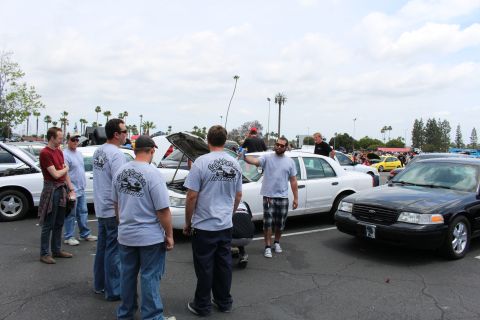 Men in gray T-shirts around a white sedan with open hood next to a black sedan
