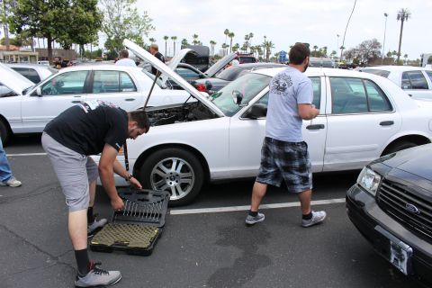 Man bent over a tool case beside a white sedan with open hood at a car meet