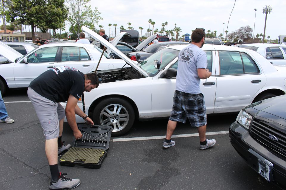 Man bent over a tool case beside a white sedan with open hood at a car meet