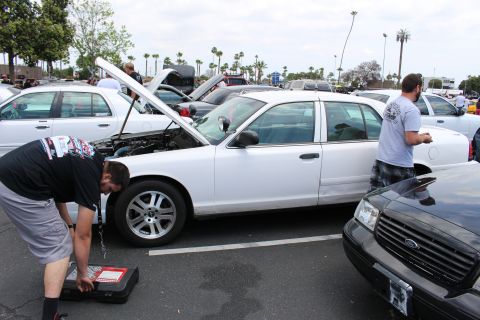 Man crouched with a tool case beside a white sedan with raised hood next to a black Ford