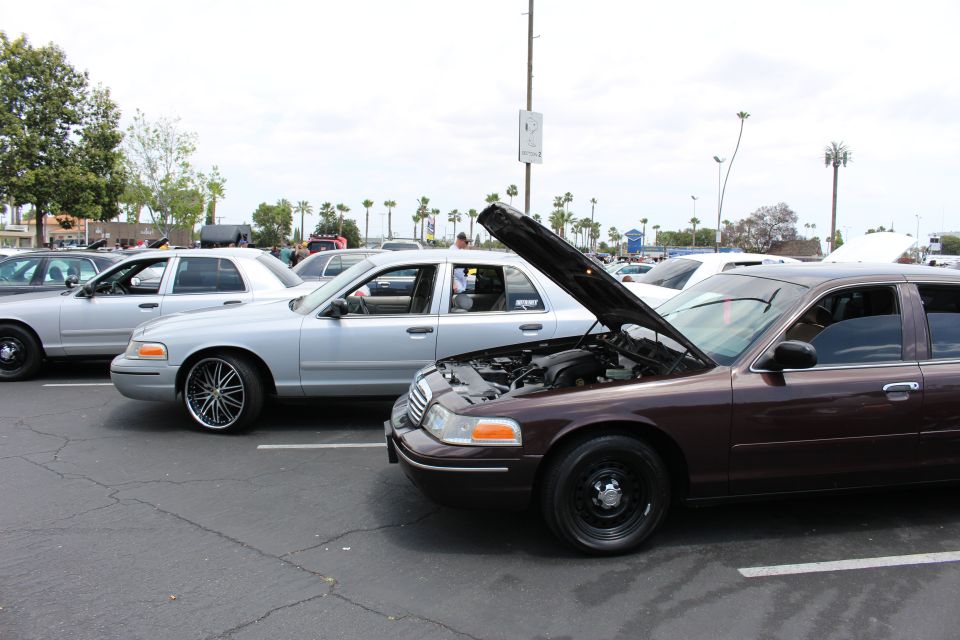 Maroon Ford sedan with open hood beside a silver sedan on chrome wheels at a meet