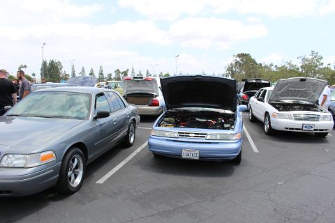 Light blue sedan with open hood flanked by Crown Victorias in a parking lot car show