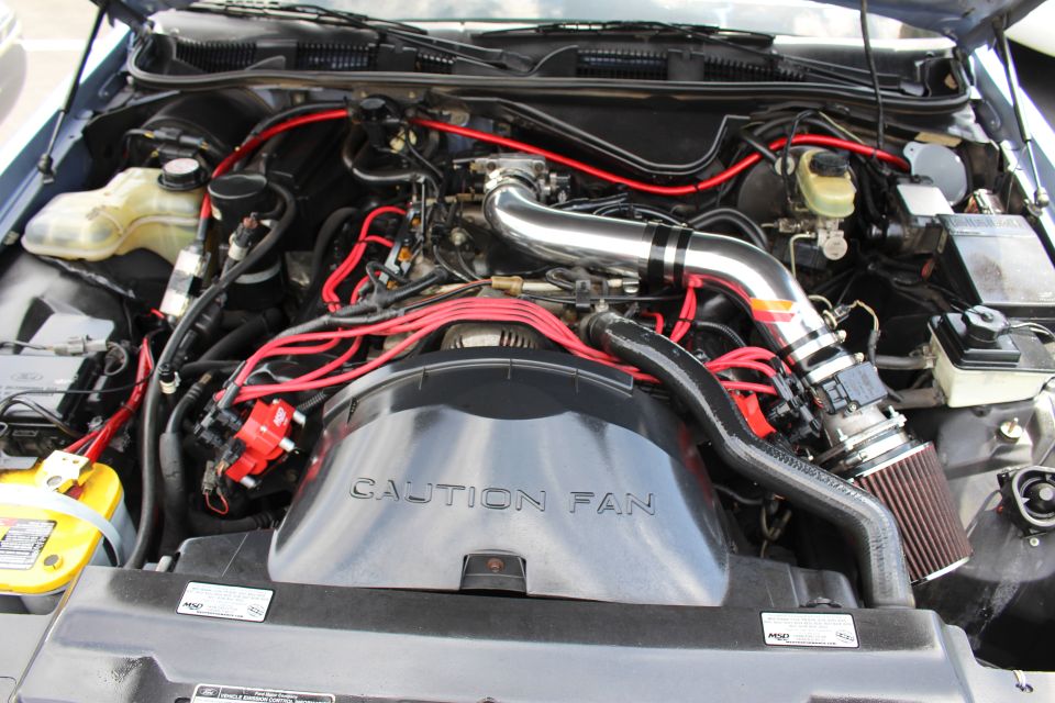 Close-up of a V8 engine bay with red wires, chrome intake and Caution Fan cover