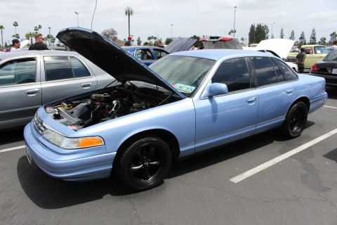 Light blue Ford Crown Victoria with black wheels and open hood at a car meet