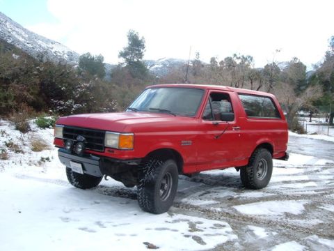 1988 Ford Bronco - This red Ford Bronco is parked in a snowy area with trees and a mountain in the background.