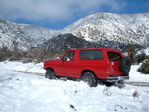 1988 Ford Bronco - A red Ford Bronco navigates a snowy mountain road, with a spare tire mounted on the rear.