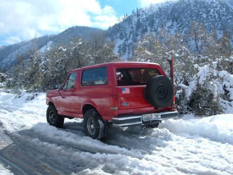 1988 Ford Bronco - A red 1988 Ford Bronco navigates a snow-covered mountain road, its spare tire mounted on the back.