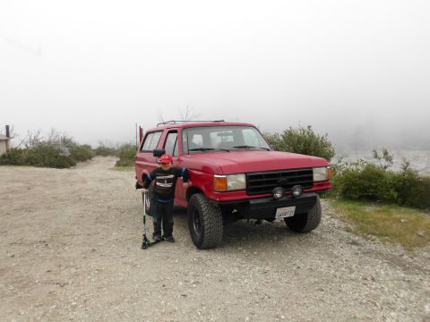 1988 Ford Bronco - A red SUV is parked on a dirt road with a boy and his scooter beside it.