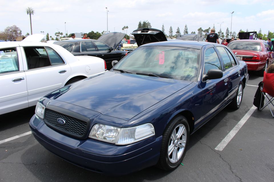 Dark blue Ford Crown Victoria sedan parked at a car show with people seated nearby
