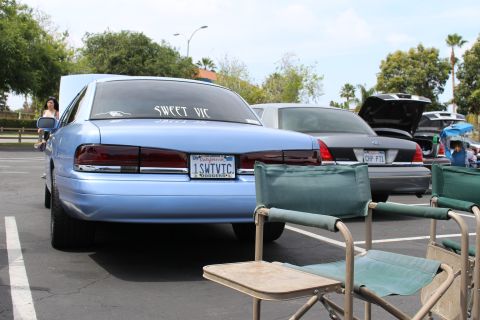 Light blue Ford sedan rear with "Sweet Vic" decal beside folding chairs at a car meet