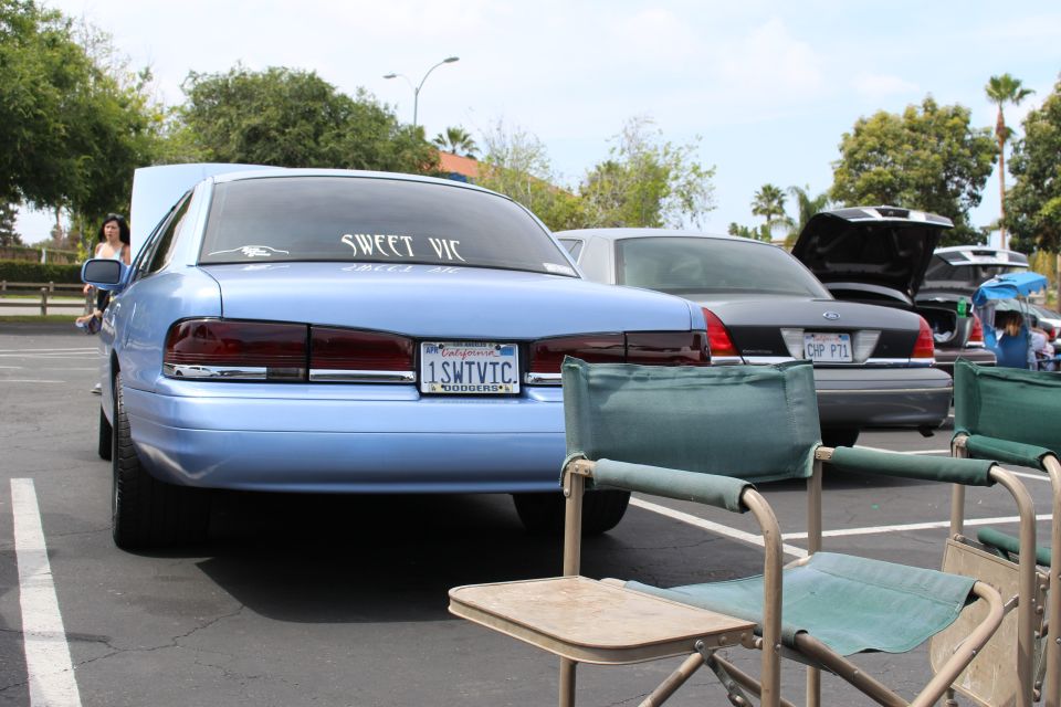 Light blue Ford sedan rear with "Sweet Vic" decal beside folding chairs at a car meet