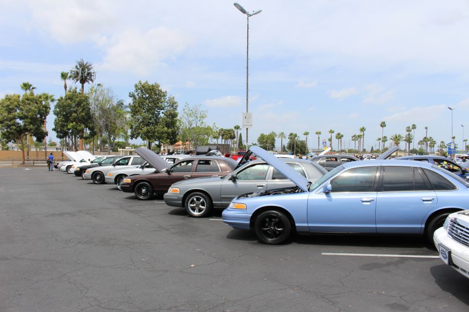 Row of sedans with open hoods at an outdoor parking lot car show, blue Ford in foreground