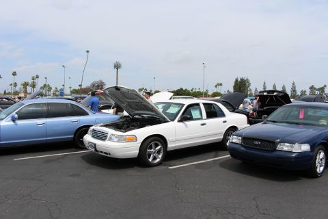 White Ford sedan with open hood between a light blue and a dark blue sedan at a car show