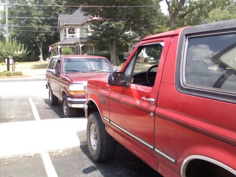 1992 Ford Bronco - Two red Ford Broncos are parked side-by-side, with the front vehicle's grille and body style indicating a 1992 model year.