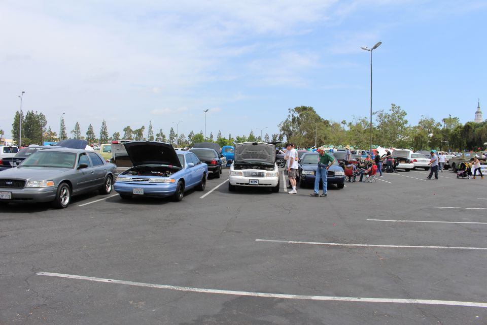 Wide view of a car show with a blue Ford and white sedan, hoods up, spectators nearby