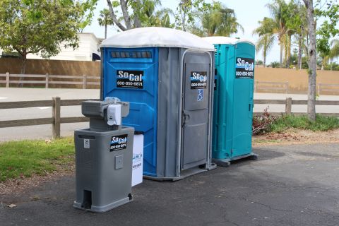 Two SoCal portable toilets and a gray hand-wash station beside a parking lot