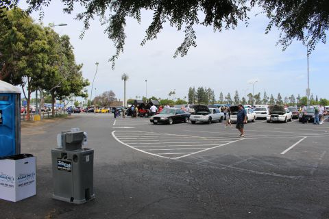 Portable toilet and trash can in foreground of a parking-lot car show