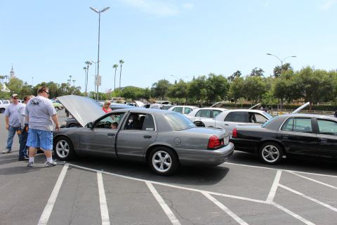 Gray sedan with hood up beside black sedan, spectators standing nearby