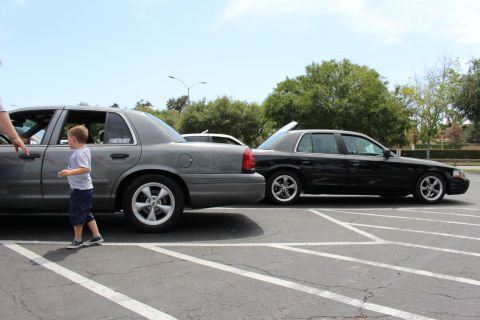 Young boy walking past a gray sedan beside a lowered black sedan in a lot