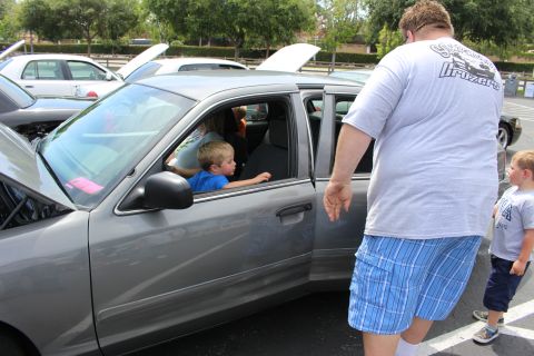 Children sitting inside a gray sedan as a large man in shorts stands by the door