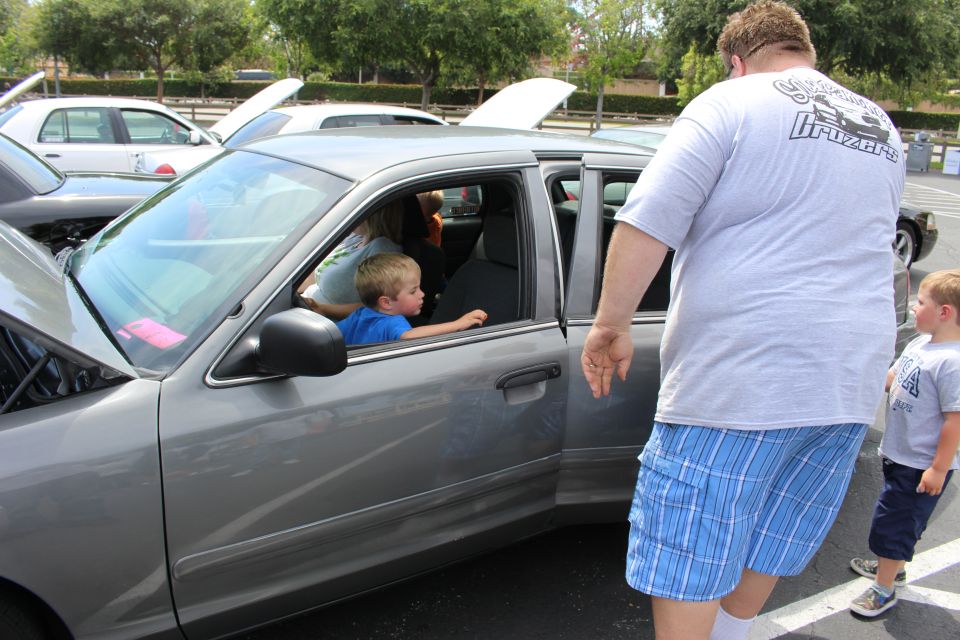 Children sitting inside a gray sedan as a large man in shorts stands by the door