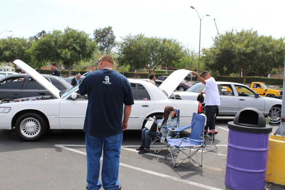 Man standing beside a white Mercury sedan with open hood at a car meet