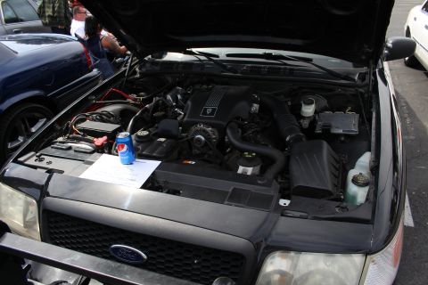 Engine bay of a black Ford sedan with hood open at a car show