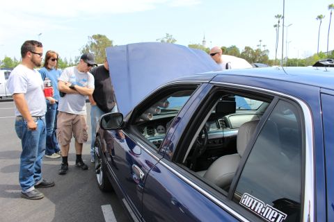 People gathered around a blue sedan with open hood at a car show
