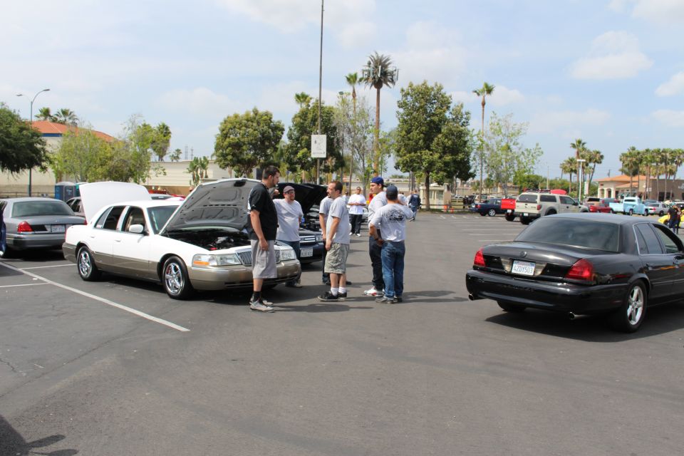 People gathered around a white sedan with open hood in a parking lot