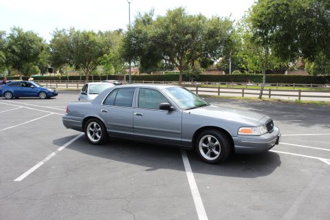 Gray full-size four-door sedan parked in an empty lot with trees and a wooden fence behind